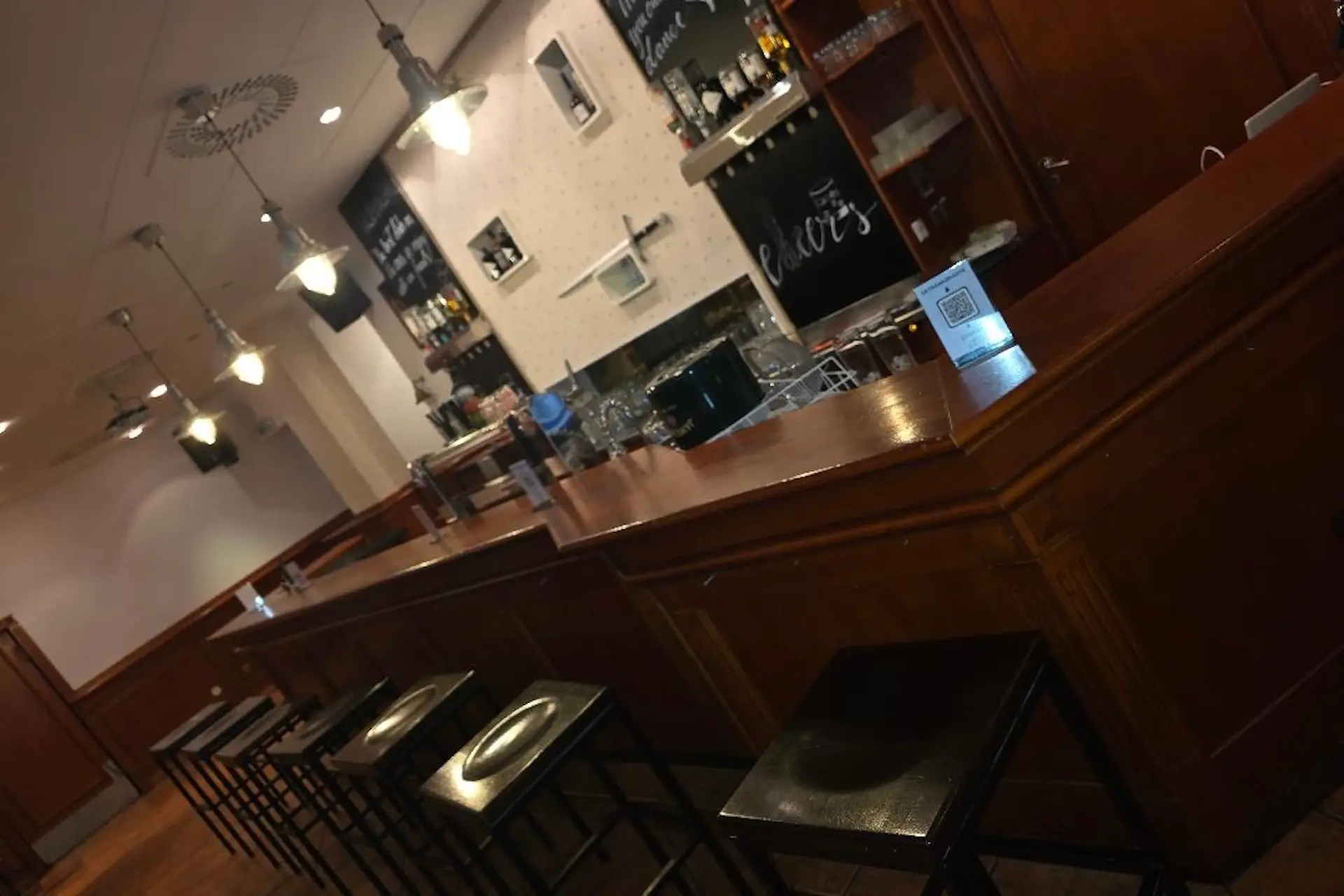 Ein hölzerner Bartresen mit hohen Hockern, Hängelampen und einem Sortiment von Flaschen und Gläsern hinter der Bar. An der Wand sind Kreidetafeln mit der Speisekarte zu sehen. Der Barbereich ist leer.