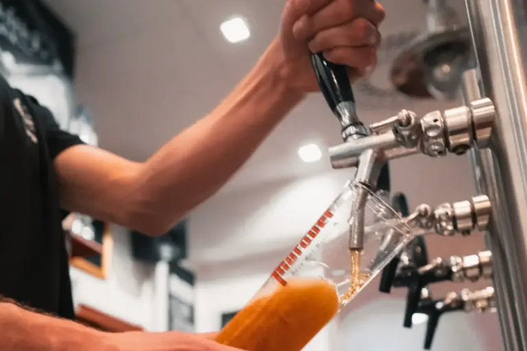 Eine Person gießt in einer Bar oder Brauerei Bier aus einem Zapfhahn in ein Glas, wobei ihre Hände und das Glas, das sich mit der bernsteinfarbenen Flüssigkeit füllt, in Großaufnahme zu sehen sind.