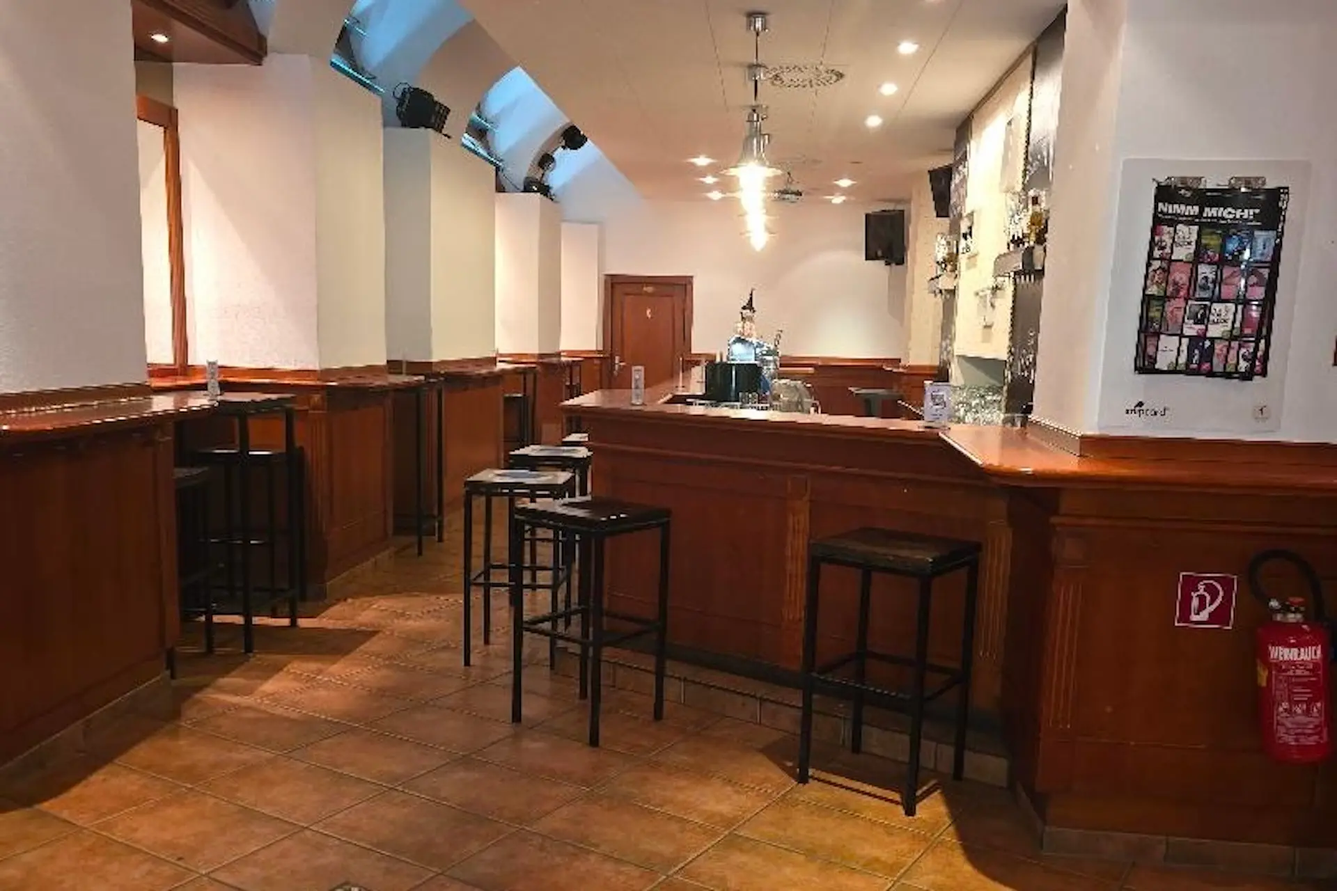 Kilimanjaro Graz Eine leere Bar mit hohen Holztresen, hohen schwarzen Hockern, gefliestem Boden und einem Feuerlöscher an der rechten Wand. Der Barbereich ist mit Flaschen und Gläsern bestückt, über denen helle Deckenleuchten hängen.