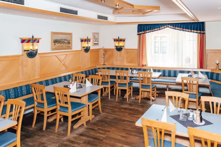 Ein heller, gemütlicher Restaurantbereich mit Holzstühlen und -tischen, blau gepolsterten Sitzbänken, gerahmten Kunstwerken an hellen Wänden und großen Fenstern mit blauen und goldenen Vorhängen, die natürliches Licht hereinlassen.