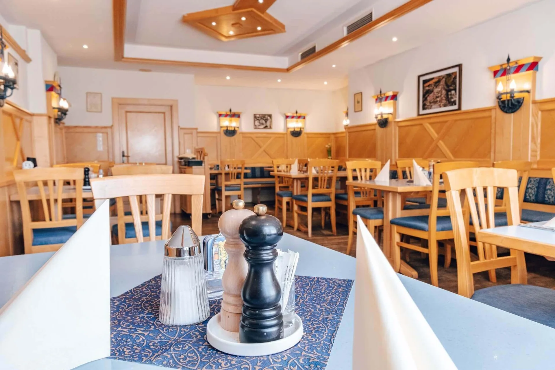 Ein heller, gemütlicher Restaurantbereich mit Holzstühlen und Tischen, die mit Servietten, Salz- und Pfefferstreuern und Gewürzen ausgestattet sind. Warme Beleuchtung und Wanddekoration schaffen eine einladende Atmosphäre.