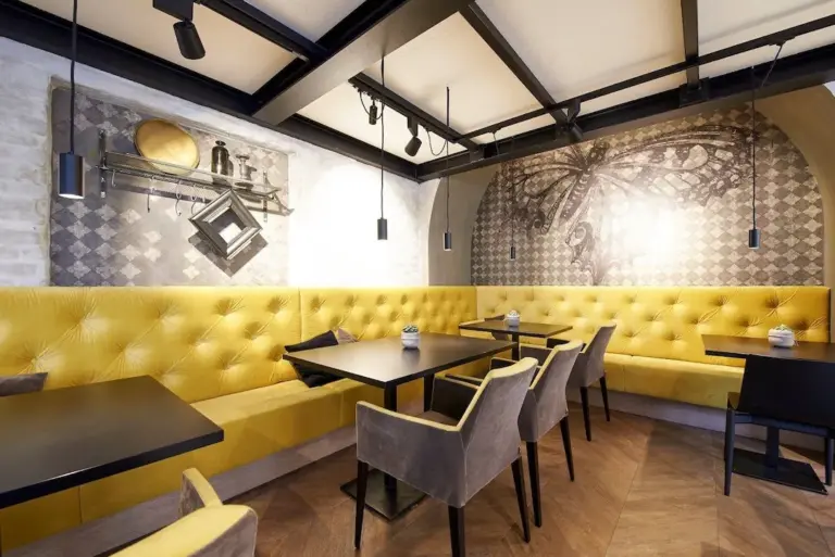 Modernes Café-Interieur mit gelben, getufteten Sitzgelegenheiten, grauen Stühlen, schwarzen Tischen, Pendelleuchten und Wandkunst mit einem großen Schmetterling und geometrischen Formen. Der Boden ist aus Holz und schafft eine warme Atmosphäre.