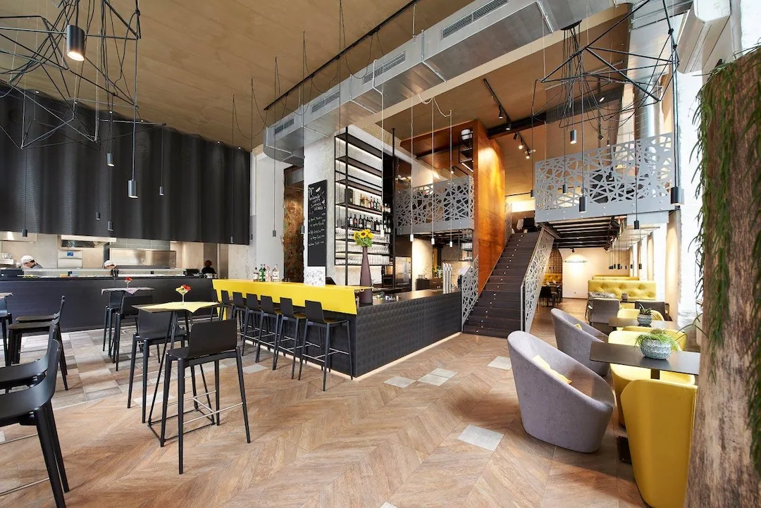 Modernes Restaurantinterieur mit hohen Decken, großen Fenstern und zeitgenössischem Dekor. Mit einer Bar mit gelben Hockern, Esstischen, grauen Sesseln und einer offenen Treppe, die zu einem Mezzanin-Sitzbereich führt.