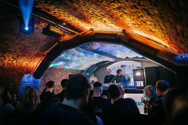 Ein Publikum genießt die Musik in einem schwach beleuchteten Underground-Club mit Backsteinwänden und gewölbter Decke, während zwei DJs in einer Kabine bei dramatischer Beleuchtung auflegen.
