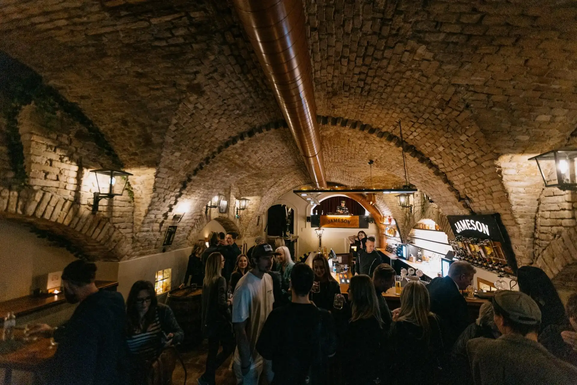 In einer gemütlichen, gewölbten Kellerbar mit warmer Beleuchtung versammelt sich ein lebhaftes Publikum. In der Nähe der Bar, wo die Drinks in rustikalem Ambiente und mit einem auffälligen Jameson-Schild serviert werden, kommen die Leute ins Gespräch.