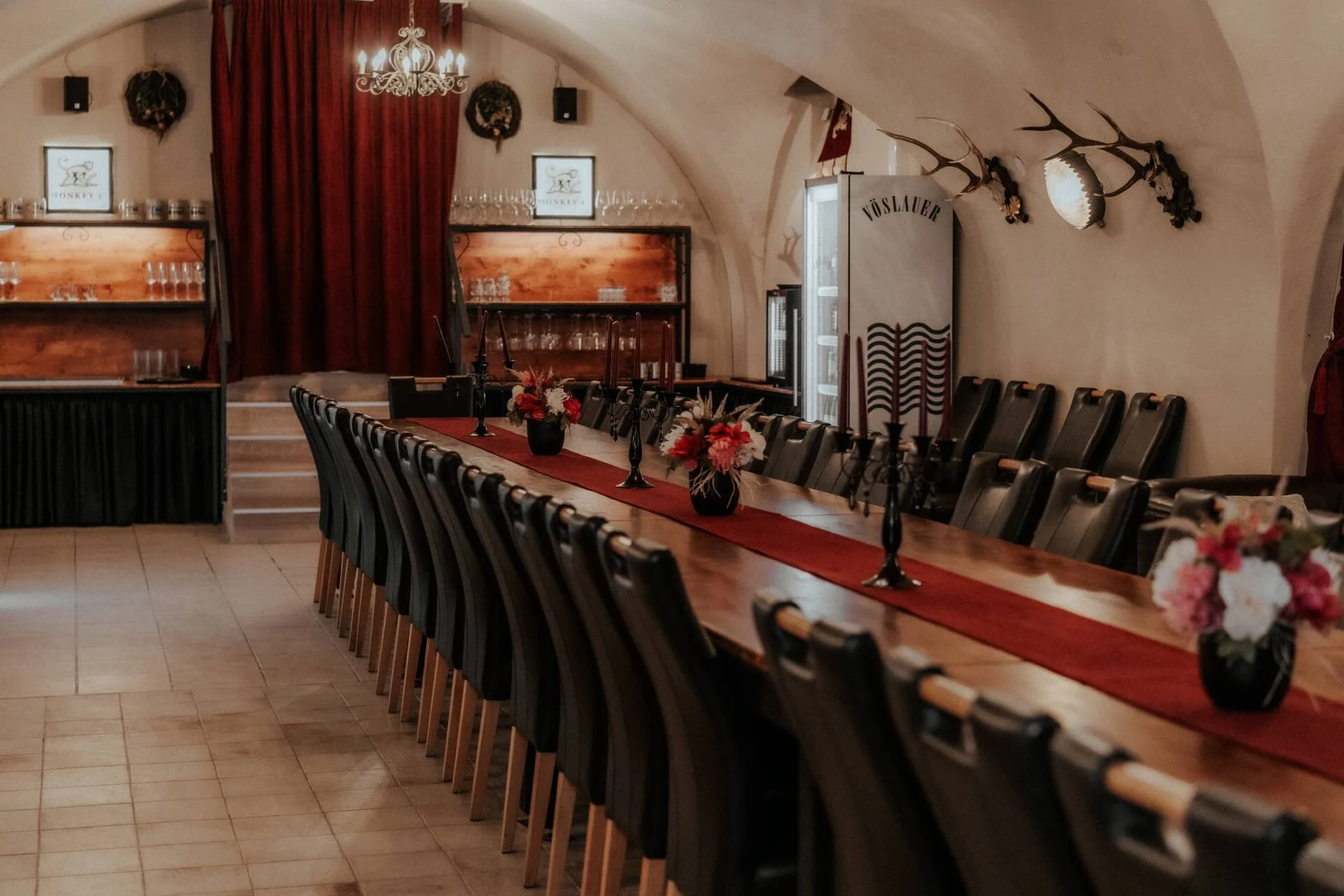 Ein langer Esstisch mit schwarzen Stühlen, roten Läufern, Kerzen und Blumenarrangements in einem eleganten Raum mit gewölbten Decken, Kronleuchtern und einem Geweih an der Wand.