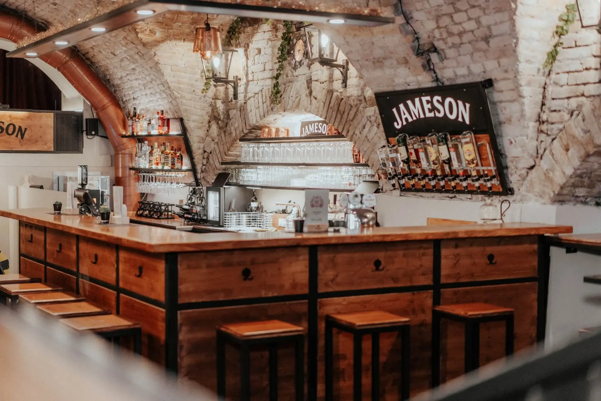 Eine gemütliche Bar mit einem hölzernen Tresen, hohen Hockern, einer gewölbten Decke aus Backstein, Regalen mit Schnapsflaschen, Hängelampen und Jameson-Schildern an den Wänden.