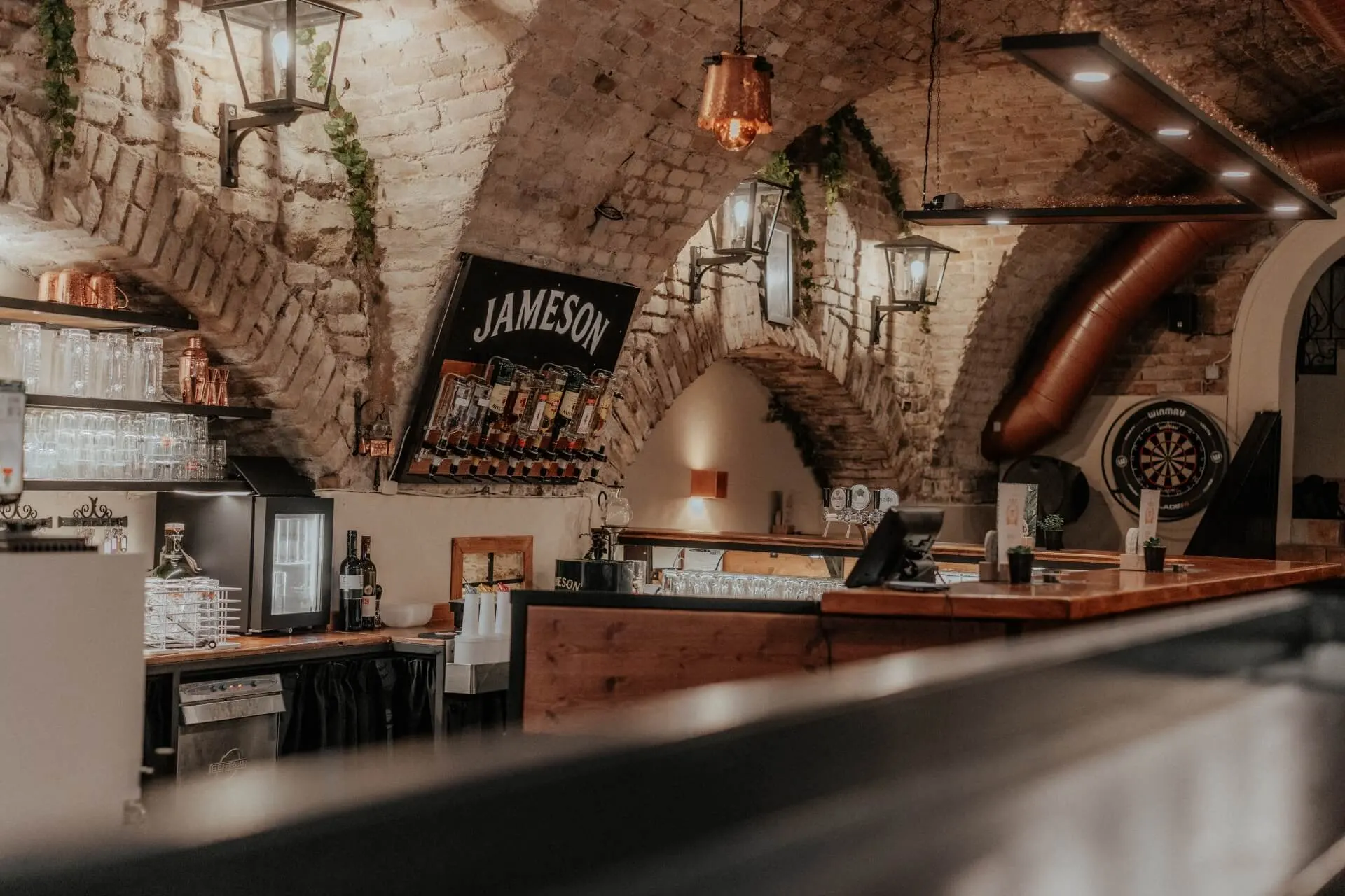 Kupferdachl Eine gemütliche Bar mit gewölbten Ziegeldecken, hängenden Laternen, einer Jameson-Whiskey-Auslage und einem Holztresen. Die Atmosphäre ist warm und einladend, die Flaschen und Gläser stehen ordentlich in den Regalen.