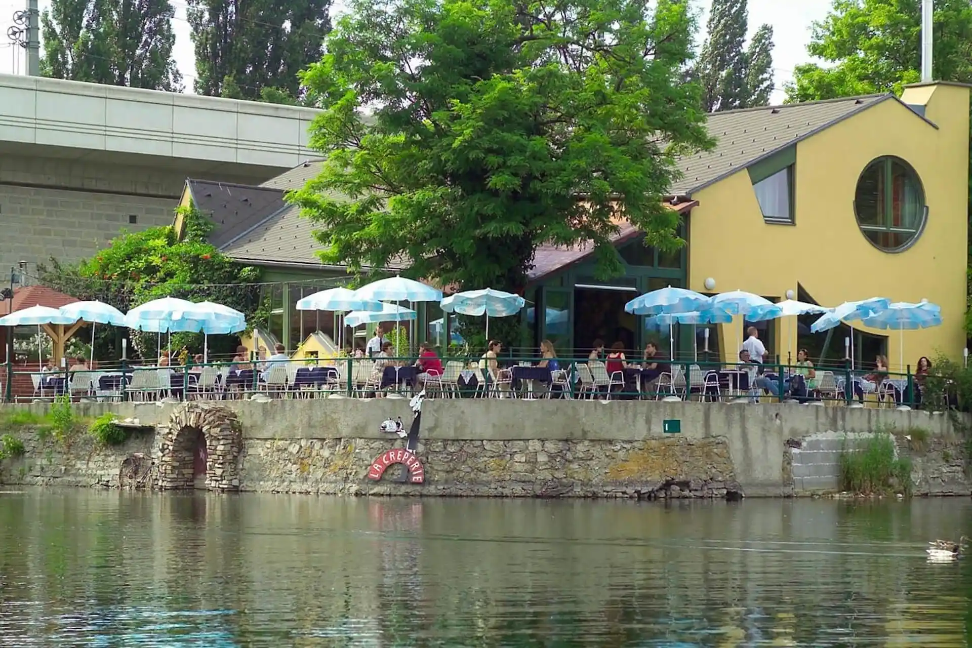Eine Restaurantterrasse mit blau-weiß gestreiften Sonnenschirmen und Blick auf einen Fluss, an deren Steinmauer ein Schild mit der Aufschrift La Crêperie angebracht ist.