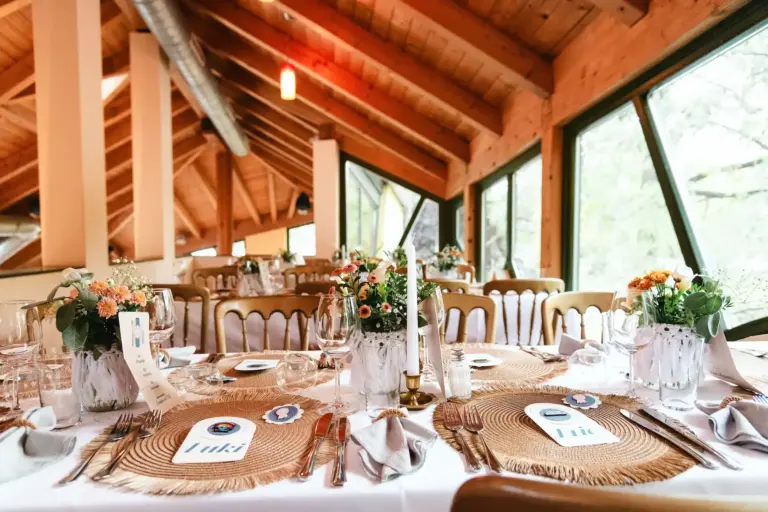 La Creperie Ein für eine Veranstaltung gedeckter Esstisch mit gewebten Tischsets, Namensschildern, Weingläsern und Blumengestecken in einem rustikalen Raum mit sichtbaren Holzbalken.