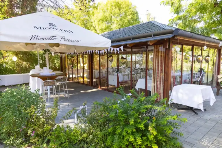 Ein Veranstaltungsbereich im Freien mit einem Mionetto Prosecco-Sonnenschirm über einem Stehtisch, direkt neben einem verglasten Pavillon, der festlich geschmückt ist.