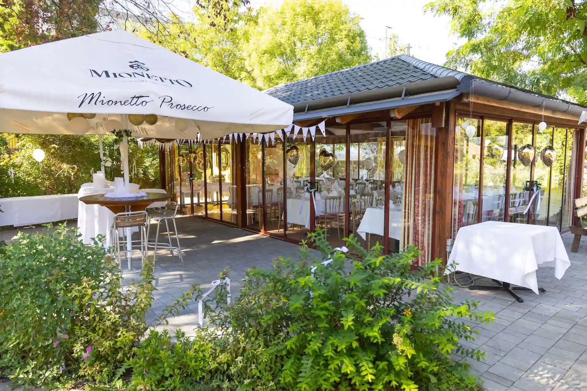 Ein Veranstaltungsbereich im Freien mit einem Mionetto Prosecco-Sonnenschirm über einem Stehtisch, direkt neben einem verglasten Pavillon, der festlich geschmückt ist.