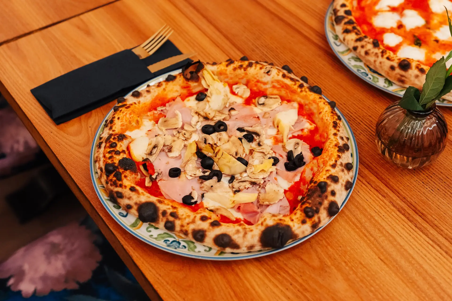 Eine im Holzofen gebackene Pizza mit Schinken, Champignons, Artischocken und schwarzen Oliven auf einem Holztisch.