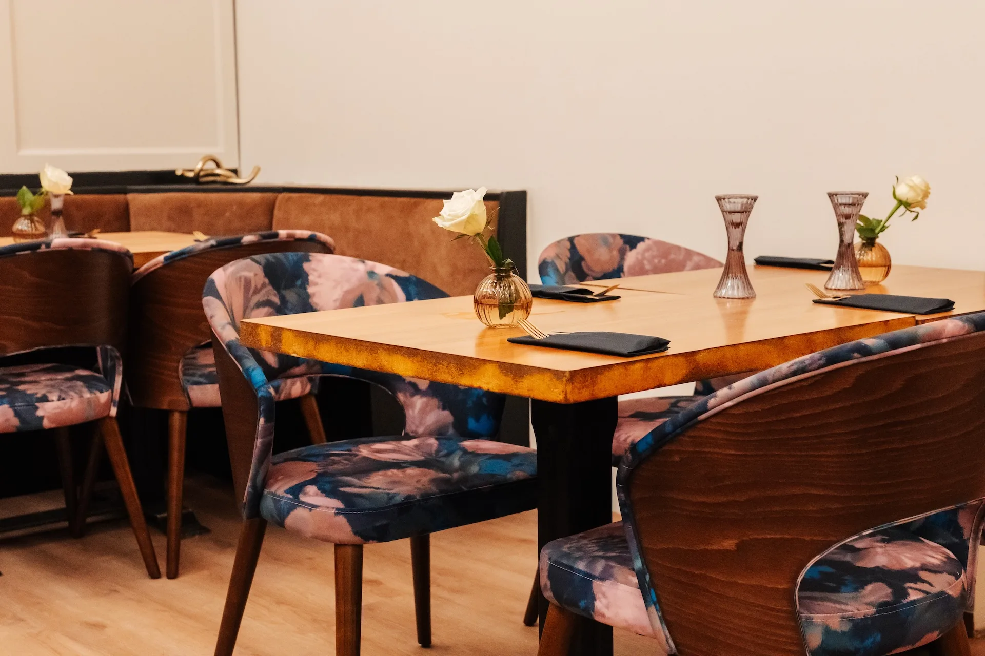 Holztische in einem Restaurant mit Stühlen, die mit blauen und rosa Blumenmustern bezogen sind; jeder Platz ist mit einer einzelnen weißen Rose in einer kleinen Glasvase geschmückt.