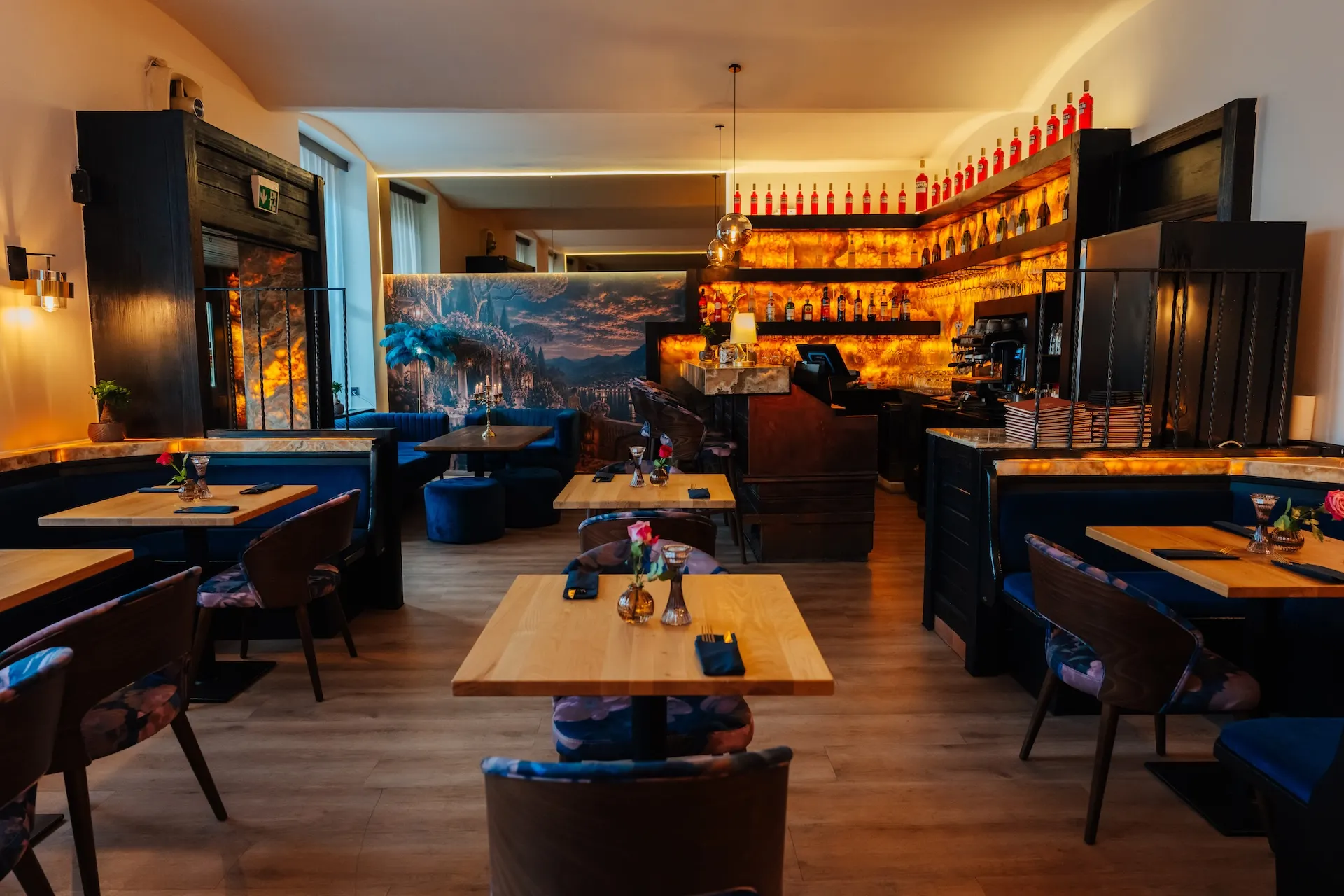 Innenansicht eines Restaurants mit Holztischen, dunkel gepolsterten Stühlen und einem Barbereich, der von warm leuchtenden Regalen beleuchtet wird.