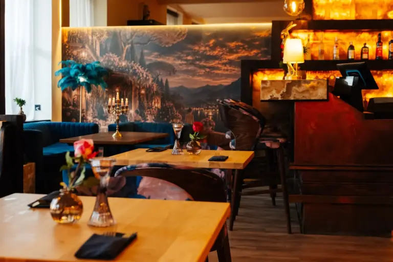 Innenansicht eines Restaurants mit Holztischen, auf denen jeweils eine einzelne Rose in einer Vase steht, vor dem Hintergrund eines Wandgemäldes, das eine Landschaft bei Sonnenuntergang zeigt, und eines hell erleuchteten Barbereichs.