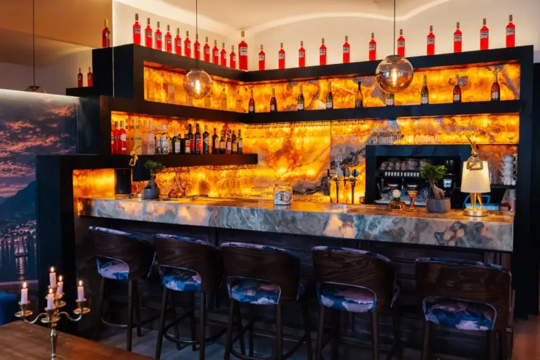 La Veneziana 2.0 Eine moderne Bar mit hinterleuchteten Steintheken, Regalen voller Spirituosenflaschen und fünf gepolsterten Barhockern aus Holz.