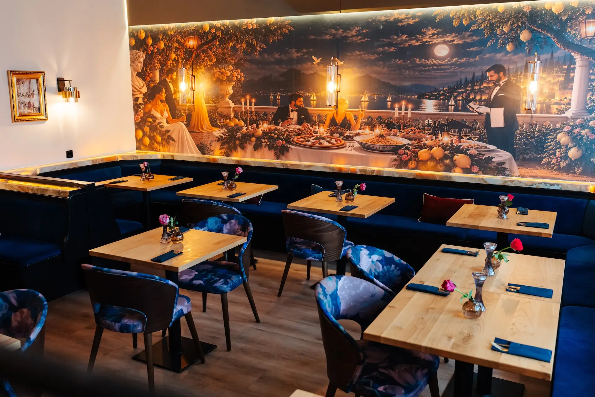 Innenraum eines Restaurants mit Holztischen, blauen Stühlen mit Blumenmuster und einem großen Wandgemälde an der Rückwand, das ein Paar zeigt, das bei Sonnenuntergang in einer Villa am Meer zu Abend isst.