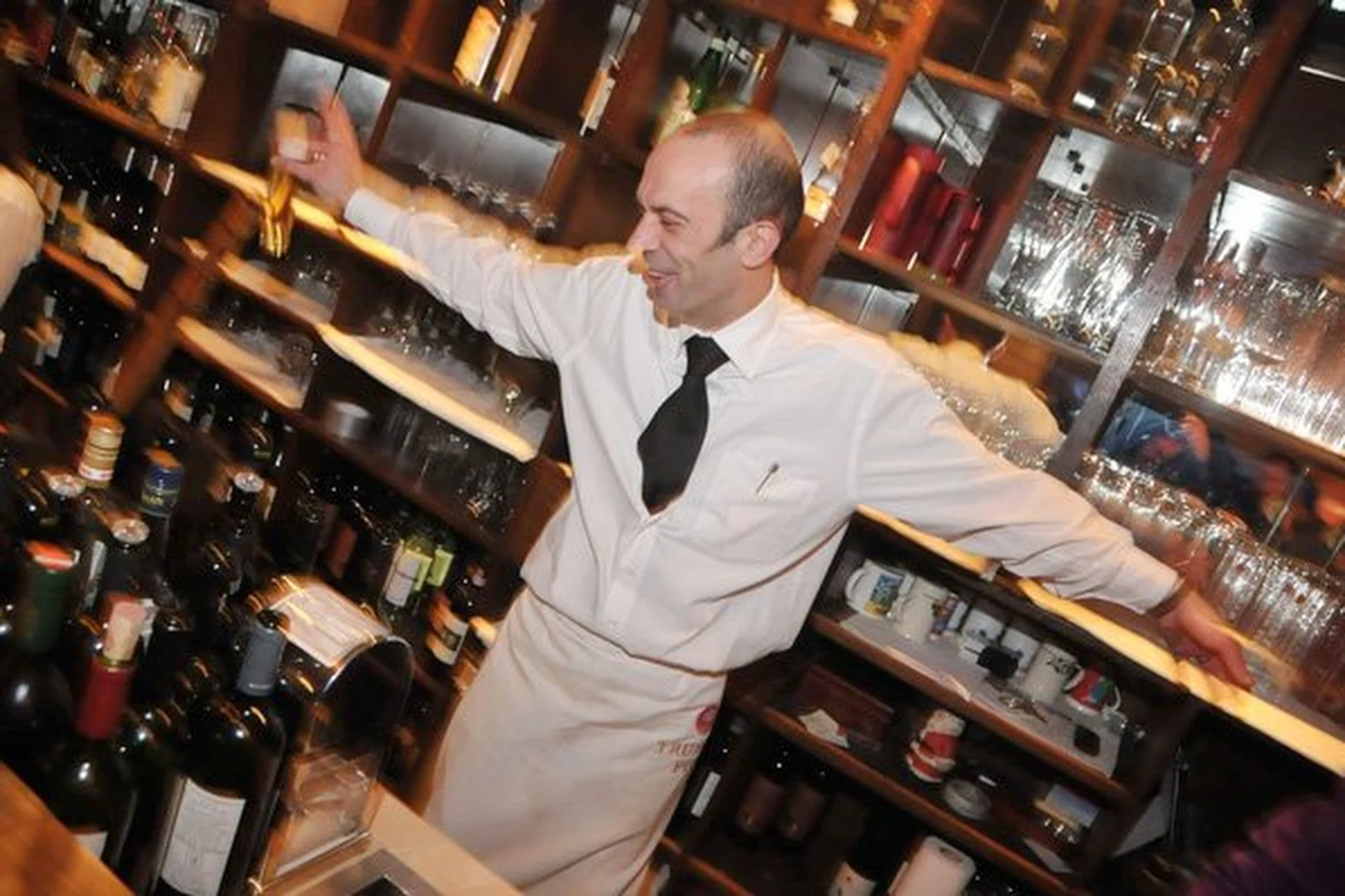 Ein lächelnder Barkeeper in weißem Hemd, schwarzer Krawatte und Schürze steht mit ausgestreckten Armen hinter einer Bar, umgeben von Flaschen und Gläsern in Regalen.