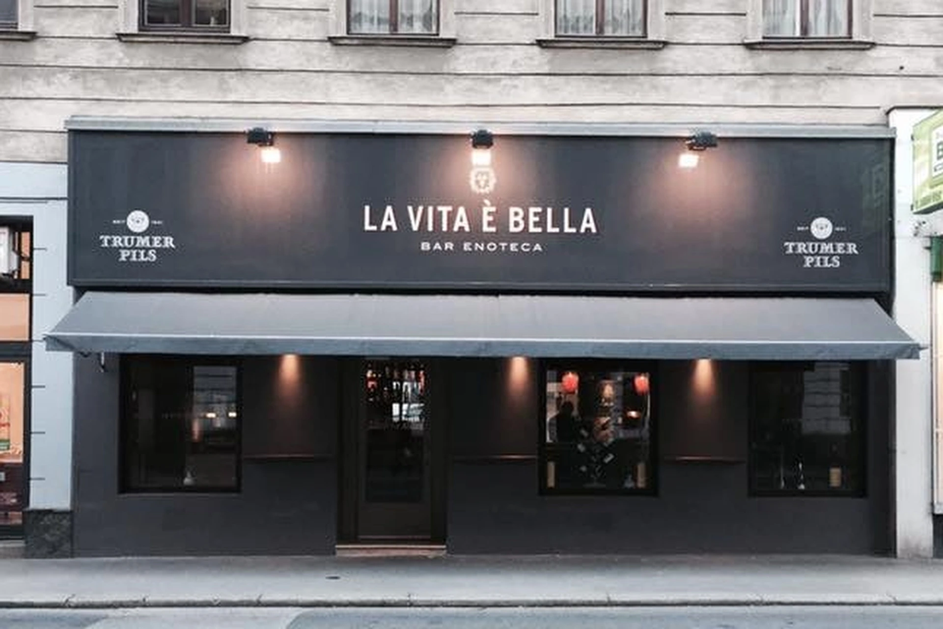 Das Äußere der La Vita È Bella Bar Enoteca, einer modernen Bar mit einer dunklen Fassade, großen Fenstern, einer Markise und einer Leuchtreklame über dem Eingang. Der Schriftzug Trumer Pils erscheint auf beiden Seiten des Schildes.