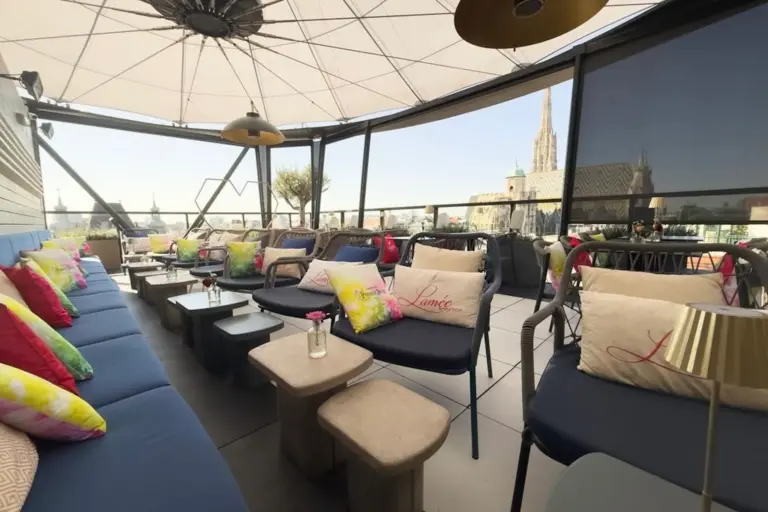 Lamée Rooftop Bar Eine Dachterrasse mit bunten Kissen auf blauen Sofas, kleinen Tischen und einem Baldachin über den Köpfen. Im Hintergrund sind unter einem klaren Himmel die Gebäude der Stadt und der Turm einer Kathedrale zu sehen.