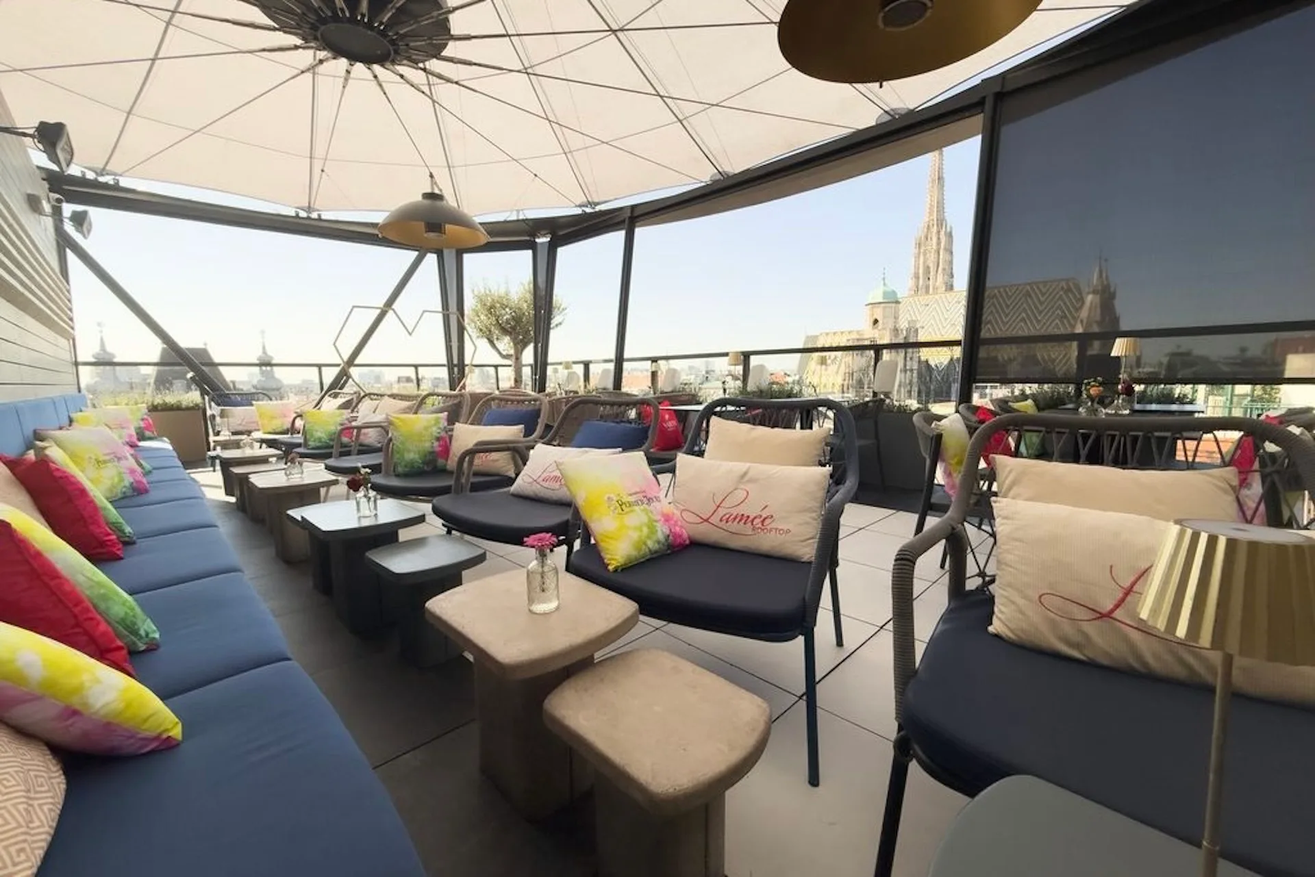 Lamée Rooftop Bar Eine Dachterrasse mit bunten Kissen auf blauen Sofas, kleinen Tischen und einem Baldachin über den Köpfen. Im Hintergrund sind unter einem klaren Himmel die Gebäude der Stadt und der Turm einer Kathedrale zu sehen.