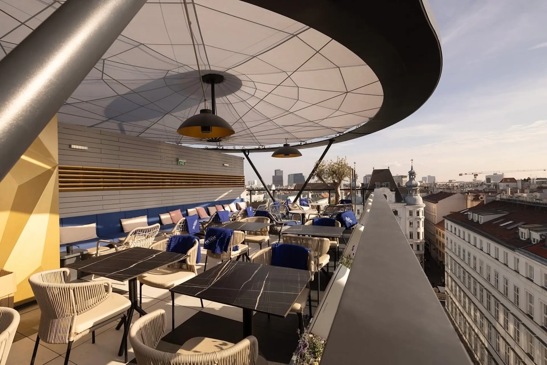 Dachterrasse mit modernen Tischen und Stühlen, beschattet von einem großen runden Sonnendach, mit Blick auf eine Stadtlandschaft mit Gebäuden unter klarem Himmel.