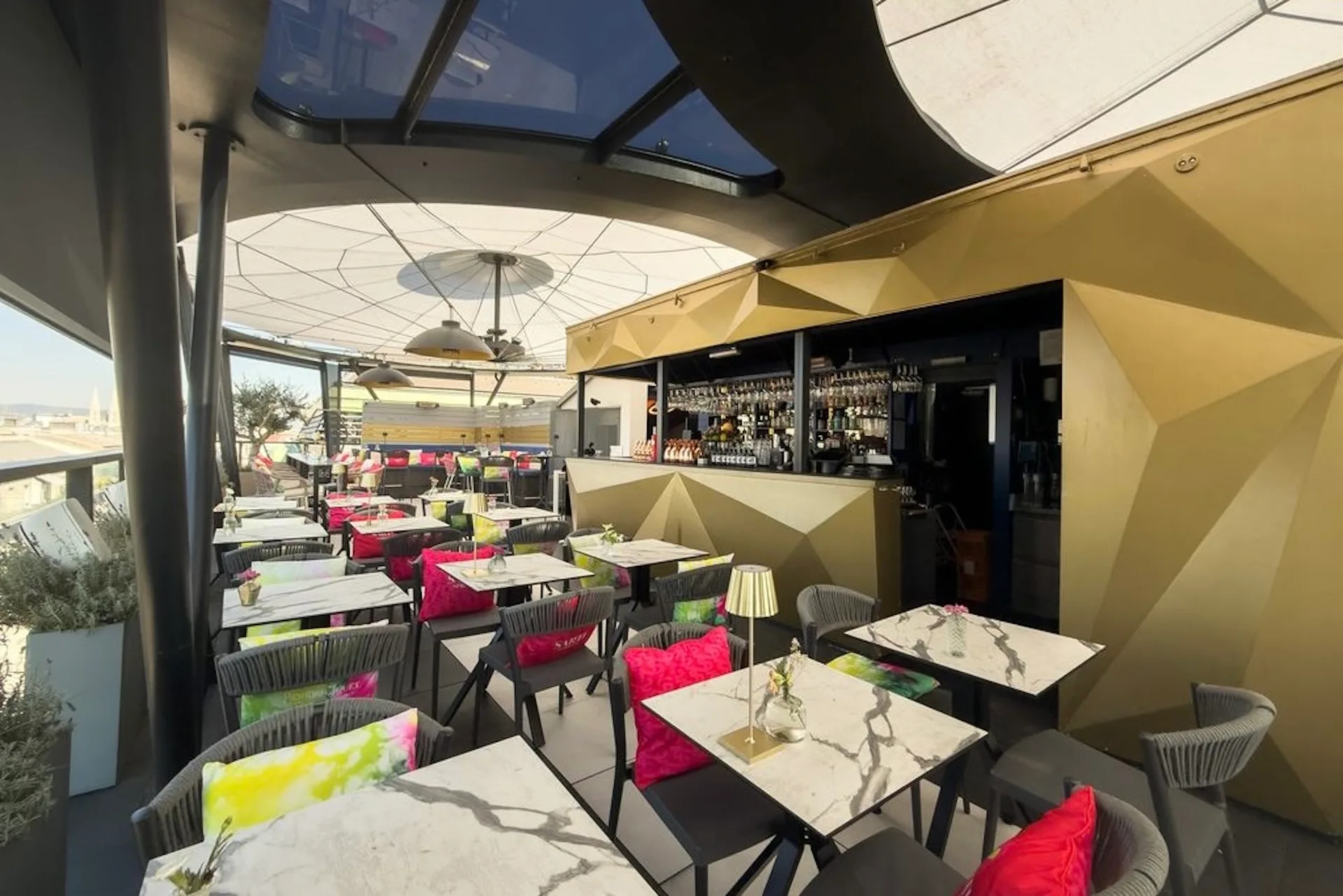 Ein Restaurant im Freien auf der Dachterrasse mit Marmortischen, bunten Kissen auf den Stühlen, einem geometrisch gestalteten, goldfarbenen Barbereich und einer teilweise verglasten Decke – und das alles unter einem strahlend blauen Himmel.
