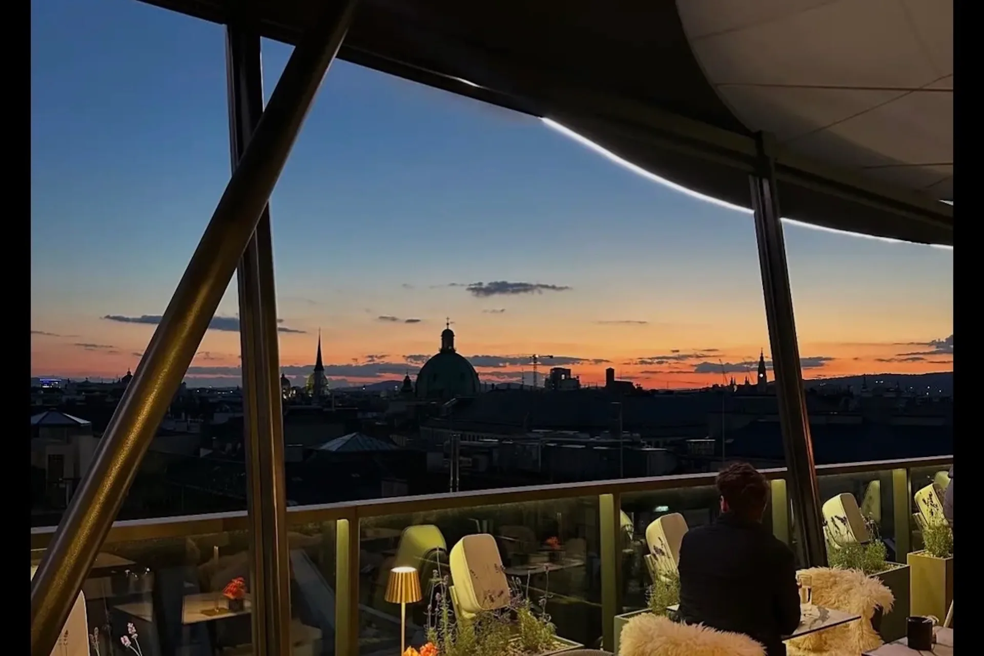 Eine Person sitzt bei Sonnenuntergang auf einer Dachterrasse und blickt auf die Skyline der Stadt mit kuppelförmigen Gebäuden und Türmen. Der Himmel ist orange und blau, und die Terrasse ist warm beleuchtet und gemütlich eingerichtet.