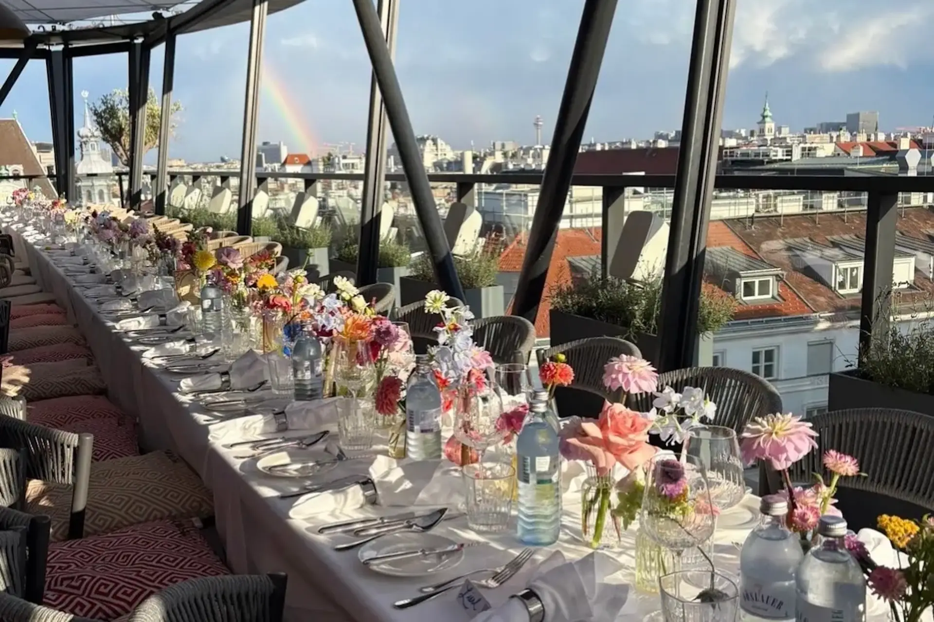 Ein langer Esstisch, gedeckt mit Gläsern, Tellern, Besteck und bunten Blumen, steht vor großen Fenstern mit Blick auf eine Stadtlandschaft, über der ein Regenbogen am Himmel zu sehen ist.