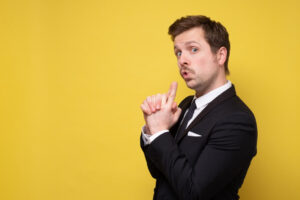 Ein Mann im Anzug posiert spielerisch vor einem gelben Hintergrund, hält seine Hände zusammen wie eine vorgetäuschte LaserTag-Pistole und macht ein überraschtes Gesicht.