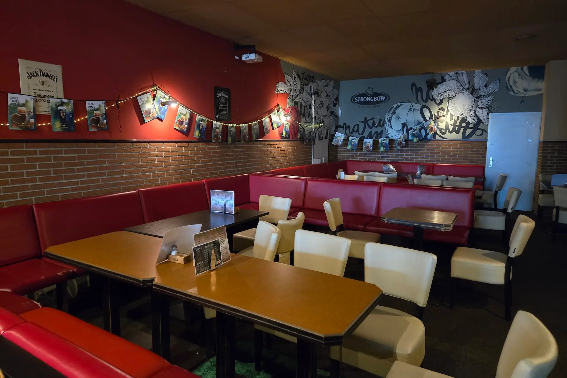 Ein gemütliches Café mit roten Ständen, weißen Stühlen und Holztischen. Die Wände sind mit Postern, Lichterketten und Graffiti-Kunst verziert. Der Raum ist leer und warm beleuchtet.