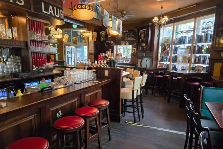 Eine gemütliche Kneipeneinrichtung mit Holzbar, rot gepolsterten Hockern, cremefarbenen Stühlen und Tischen an großen Fenstern. In den Regalen stehen Gläser und Flaschen, und die warme Beleuchtung schafft eine einladende Atmosphäre.