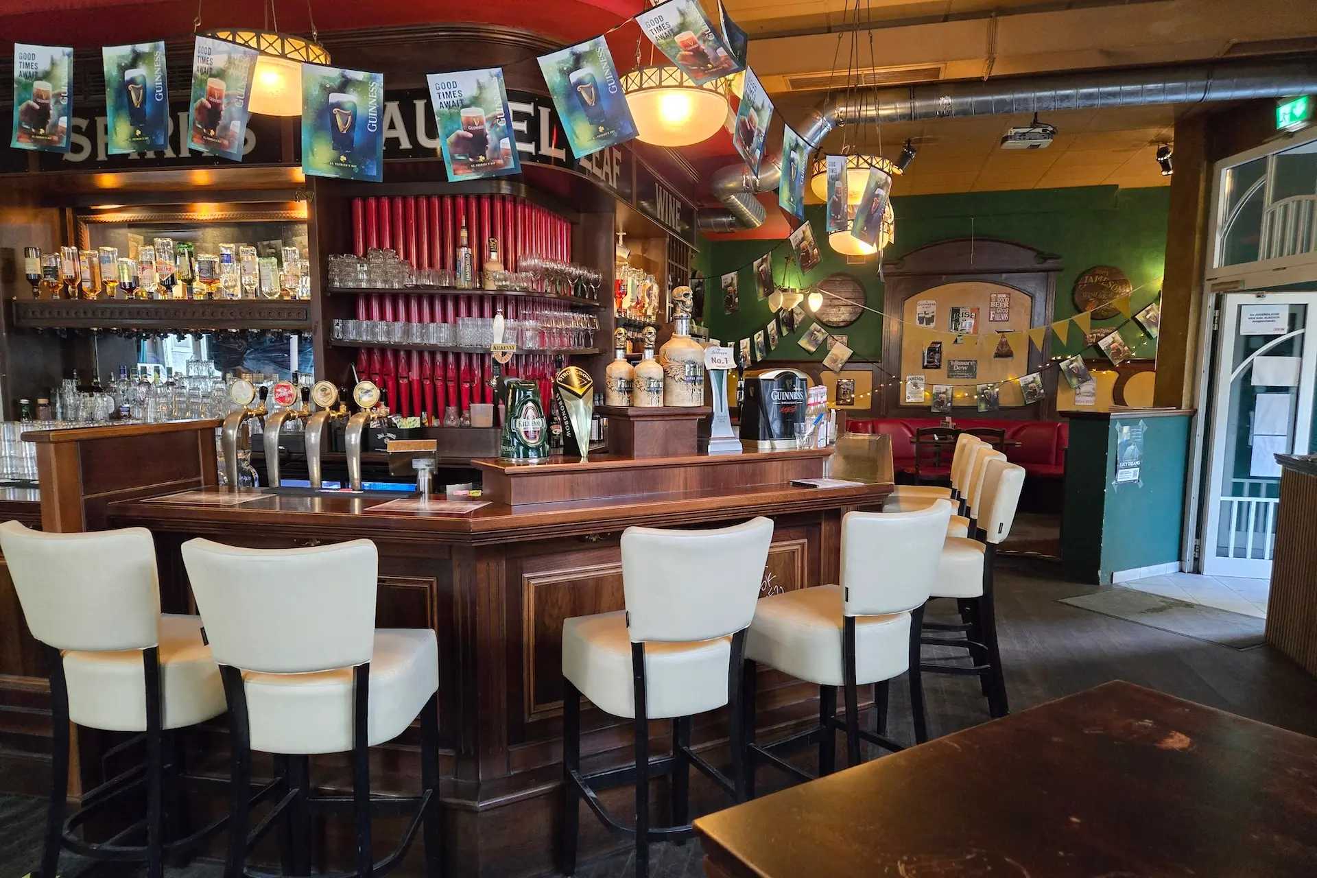 Eine gemütliche Kneipe mit einer hölzernen Theke, cremefarbenen Barhockern, Bierzapfhähnen, Regalen mit Gläsern, festlichen Bannern und warmer Beleuchtung. Die Wände sind mit Postern und verschiedenen Erinnerungsstücken geschmückt.