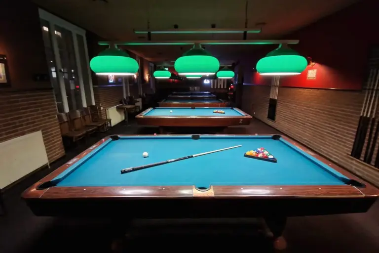 In einem schwach beleuchteten Raum stehen mehrere Billardtische mit hellgrünen Scheinwerfern übereinander. Auf dem nächstgelegenen Tisch liegen ein Queue, ein weißer Spielball und ein Satz farbiger Kugeln bereit.
