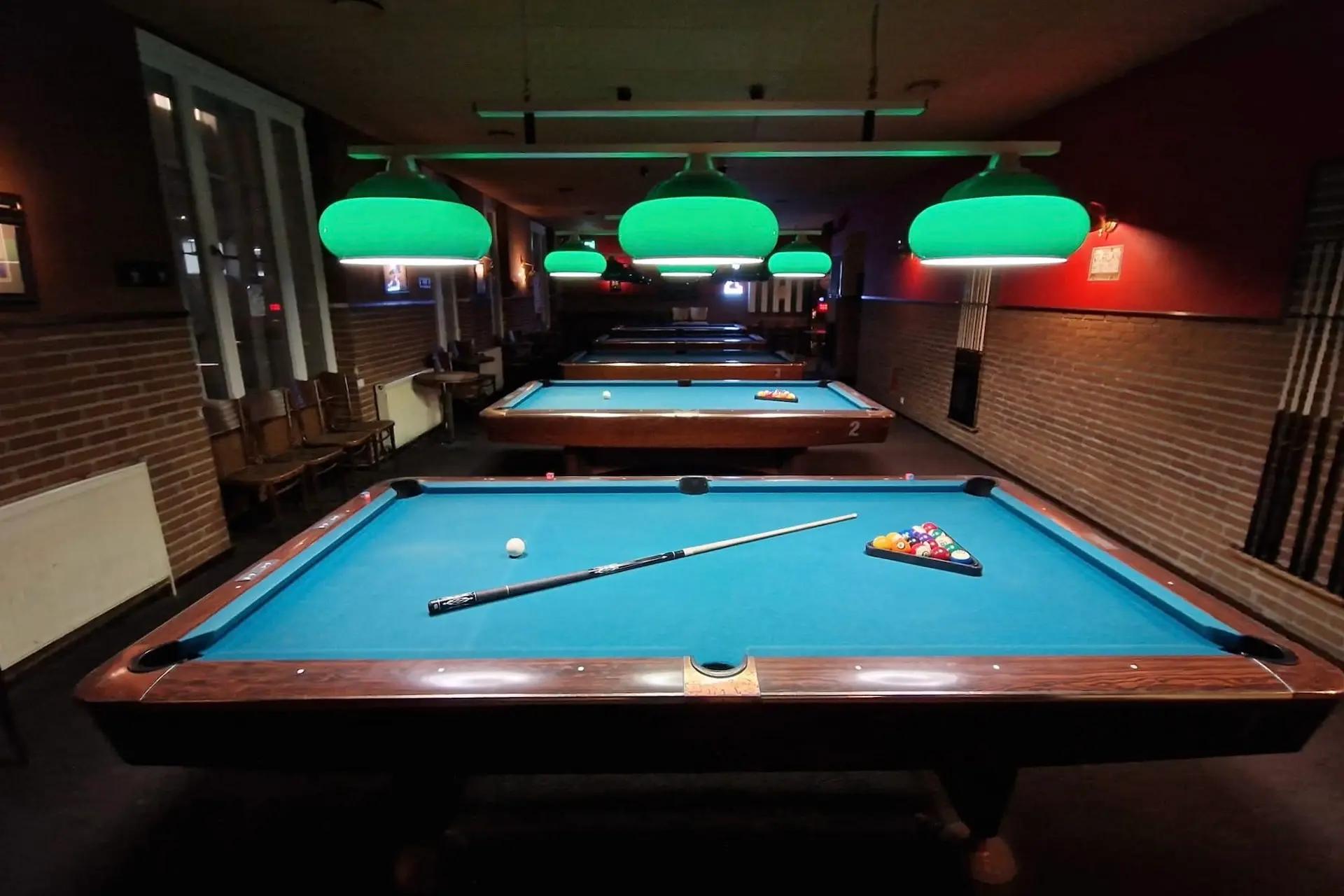 In einem schwach beleuchteten Raum stehen mehrere Billardtische mit hellgrünen Scheinwerfern übereinander. Auf dem nächstgelegenen Tisch liegen ein Queue, ein weißer Spielball und ein Satz farbiger Kugeln bereit.