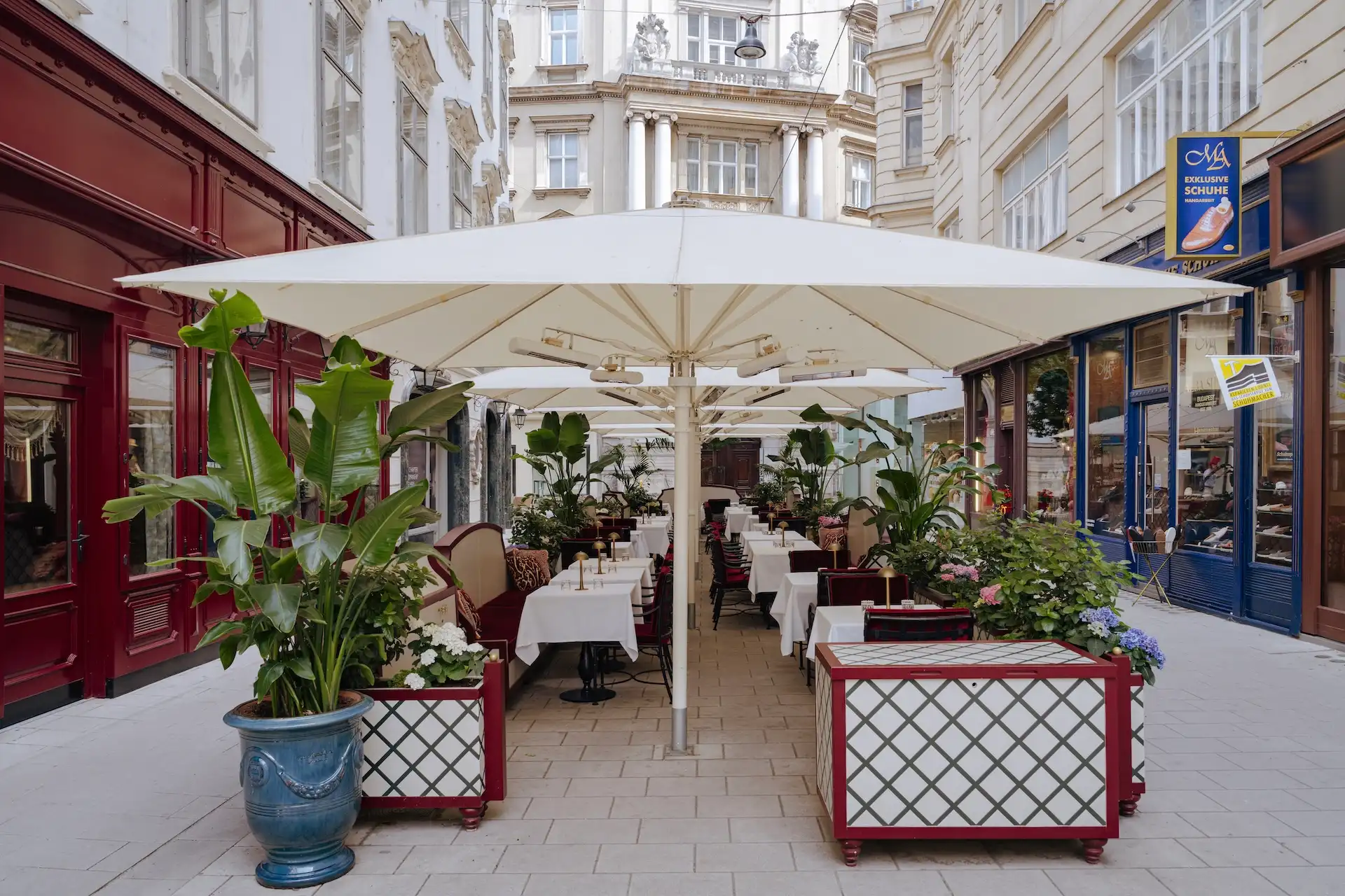 Ein Straßencafé mit weißen Sonnenschirmen, Tischen mit weißen Tischdecken und Topfpflanzen, gelegen zwischen eleganten historischen Gebäuden in einer gepflasterten, schmalen Straße.
