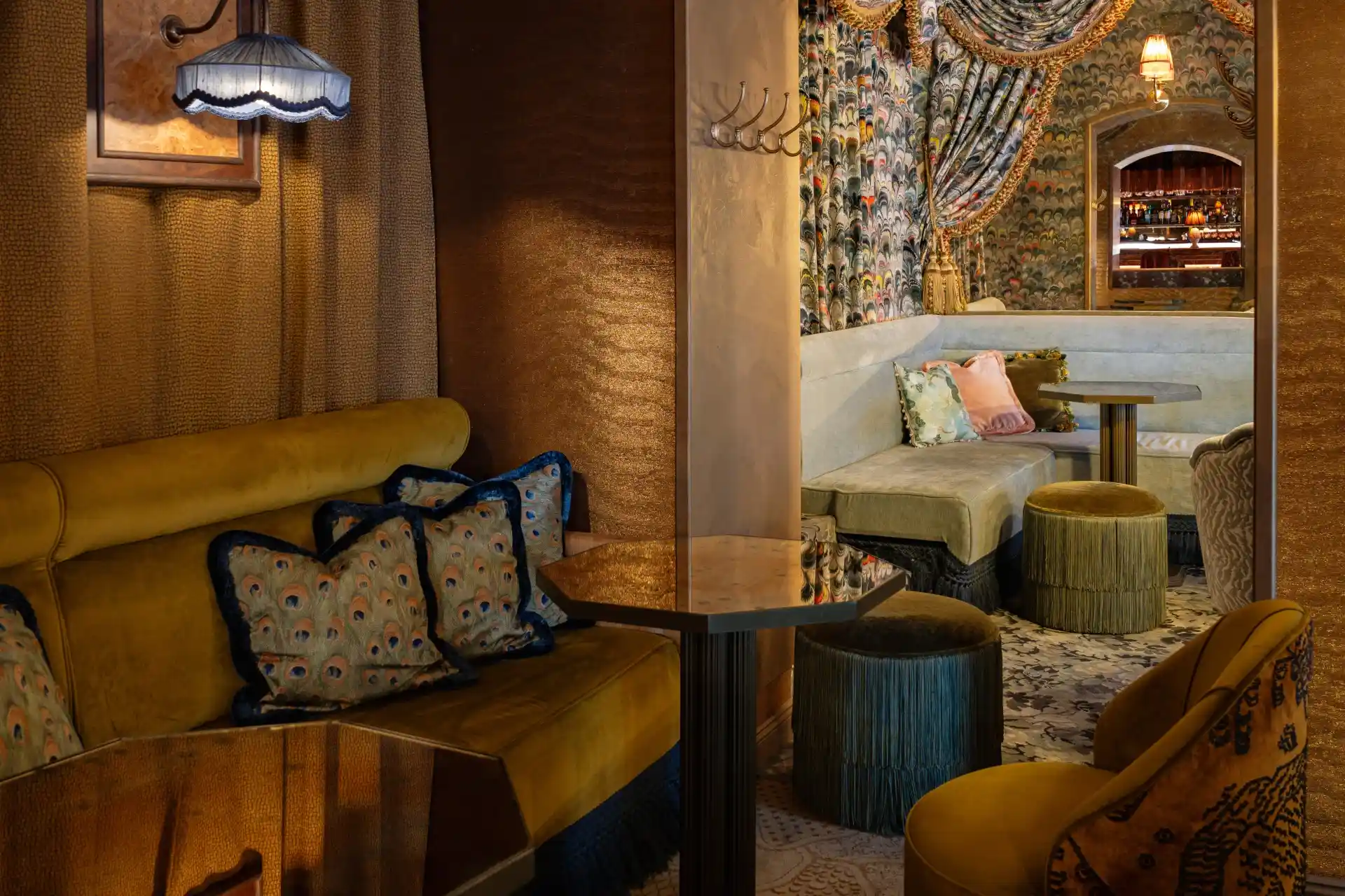 Eine gemütliche Lounge im Vintage-Stil mit gemusterten Kissen, Samtsitzmöbeln, Hockern mit Fransen, strukturierten Wänden und einem dekorativen Wandbild. Warme Beleuchtung und ein kleiner Barbereich sorgen für eine einladende, intime Atmosphäre.