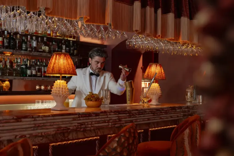 Ein Barkeeper in einer weißen Jacke mixt hinter einer Marmor-Theke einen Drink, umgeben von Gläsern, warmer Beleuchtung und eleganter Einrichtung in einer gemütlichen, stilvollen Lounge.