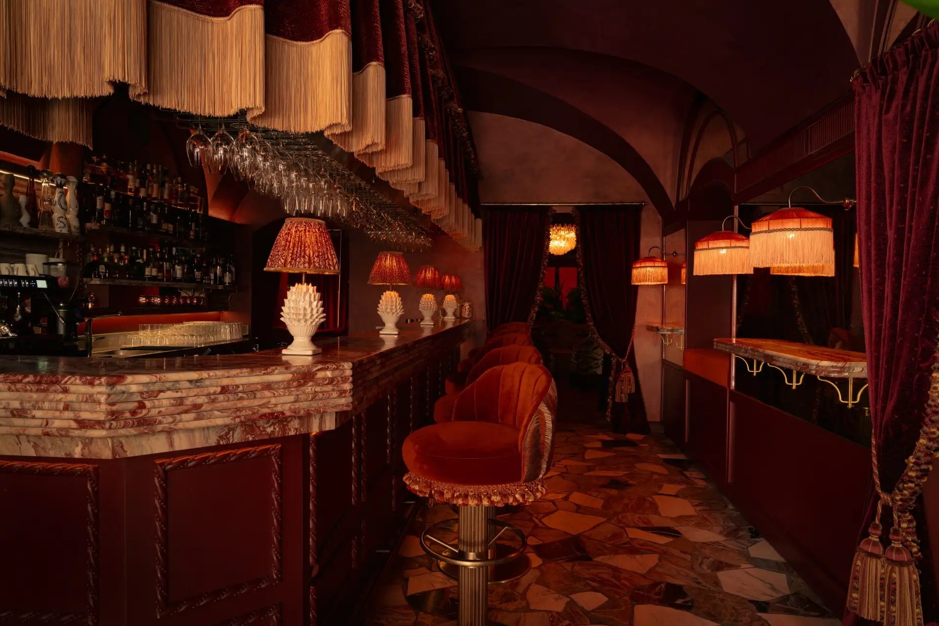 Eine schwach beleuchtete Bar mit roten Samthockern, einer Marmortheke und Vintage-Lampen mit Fransen. Der Boden besteht aus gemustertem Stein, und die Einrichtung wirkt warm und luxuriös mit satten, dunklen Farbtönen.
