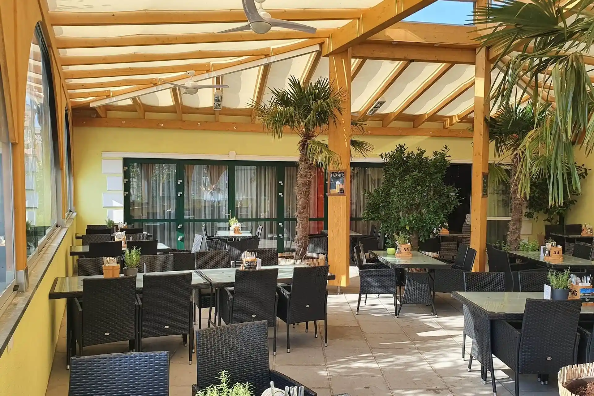 Eine überdachte Außenterrasse in einem Restaurant mit schwarzen Korbtischen und -stühlen, Topfpflanzen und großen Fenstern. Das Sonnenlicht fällt durch das hölzerne Pergoladach und schafft eine helle und einladende Atmosphäre.