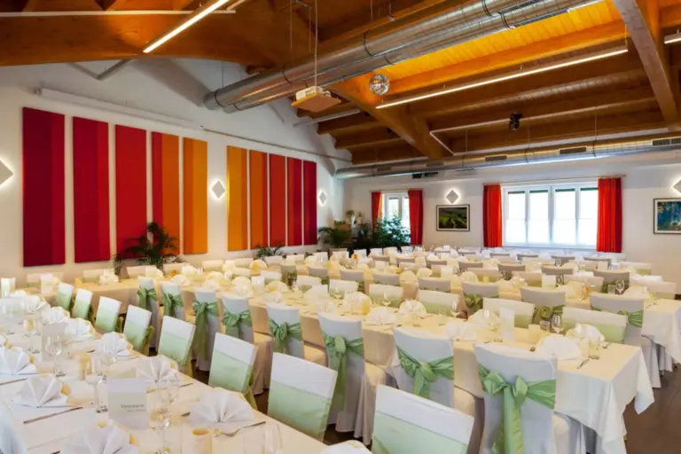 Ein großer Bankettsaal mit Tischreihen, die mit weißen Tüchern und grünen Stuhlschärpen für eine Veranstaltung gedeckt sind. Der Raum hat bunte Wandpaneele, Holzbalken und große Fenster mit roten Vorhängen.