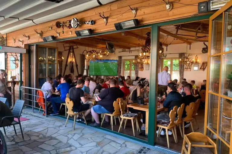 Eine Gruppe von Menschen sitzt an Holztischen in einem rustikalen Restaurant und sieht sich ein Fußballspiel auf einem an der Wand montierten Fernseher an. Die großen Fenster sind offen, und der Raum ist mit Geweihen und landwirtschaftlichen Geräten dekoriert.