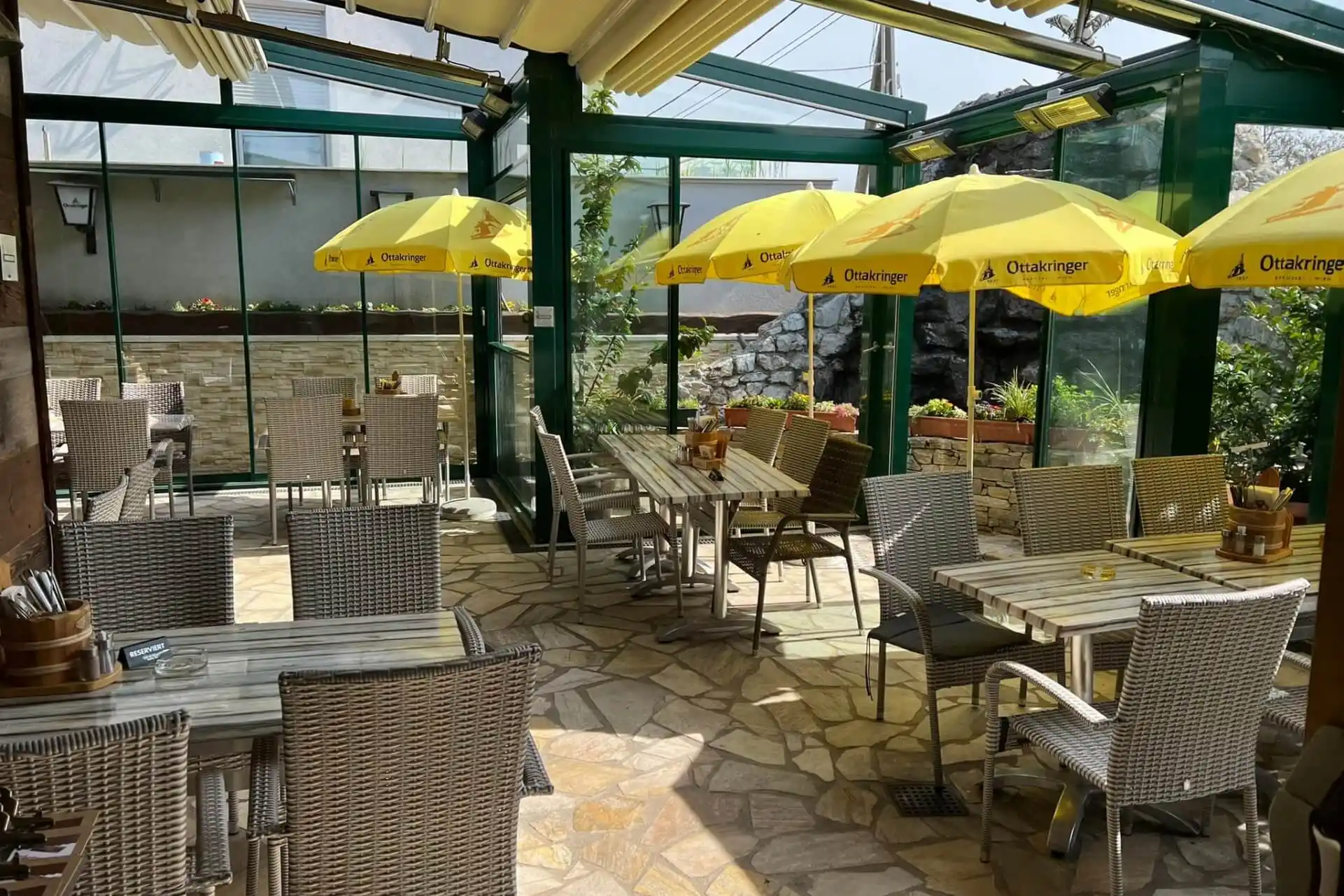 Eine helle Café-Terrasse mit Korbstühlen und Holztischen, die von gelben Sonnenschirmen beschattet werden. Das Sonnenlicht strömt durch ein Glasdach und die Wände, und Topfpflanzen säumen die Ränder. Der Raum wirkt einladend und gut gepflegt.