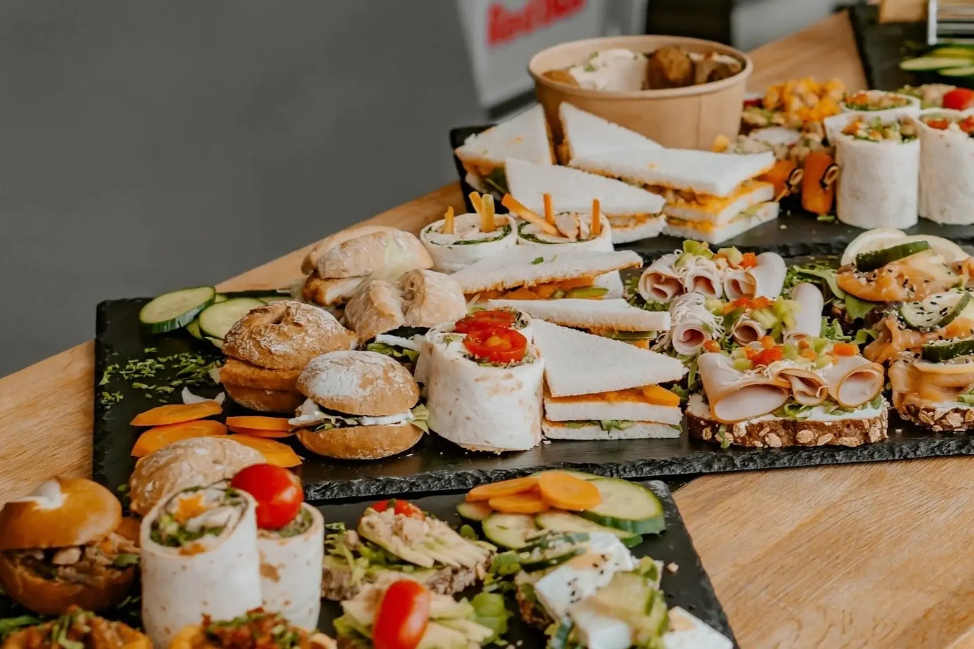 Eine Vielzahl von Fingerfood auf schwarzen Tabletts, darunter Mini-Sandwiches, Wraps, Sliders und geschnittenes Gemüse, ordentlich auf einem Holztisch angeordnet.
