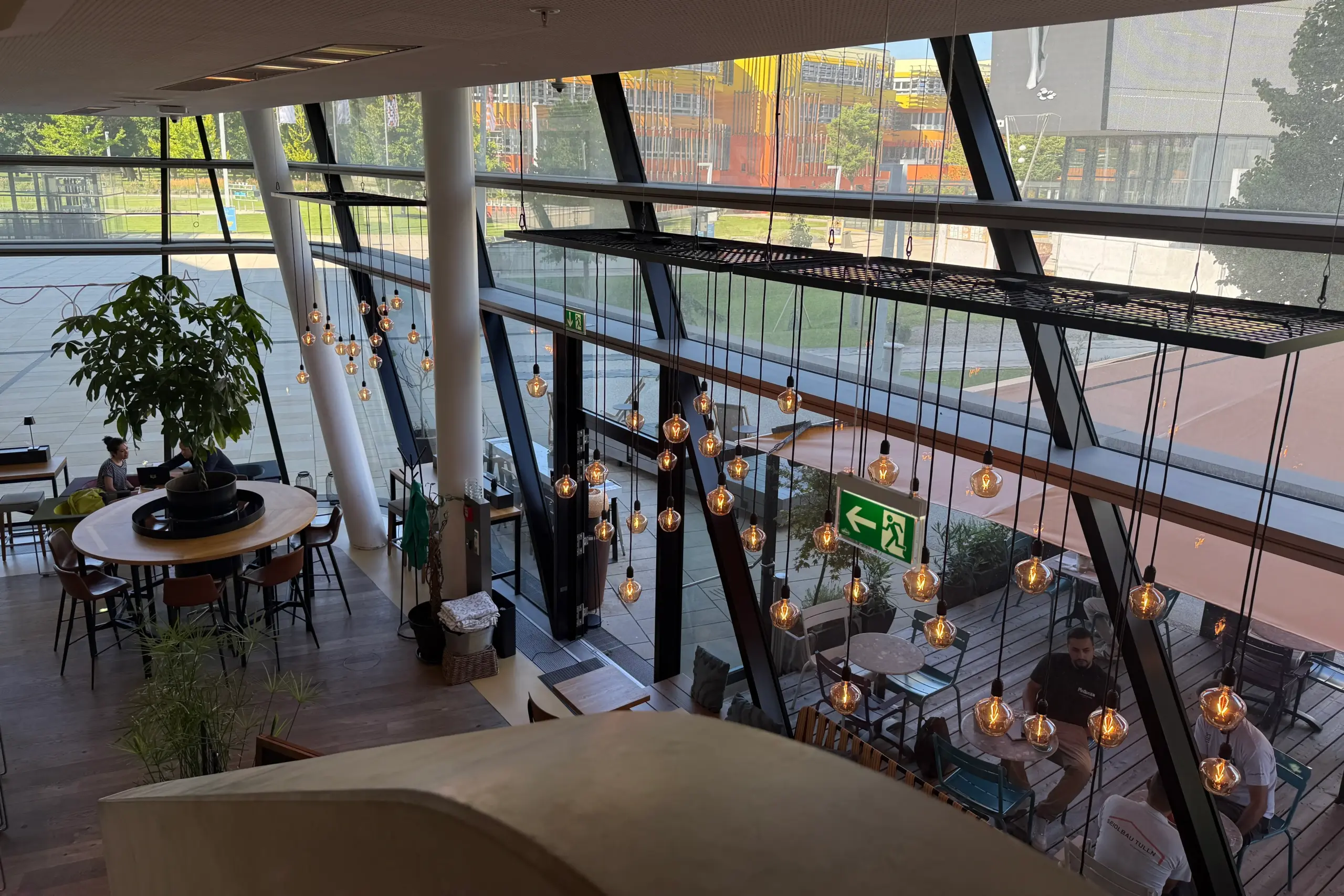 Modernes Innencafé mit großen Fenstern, hängenden Glühbirnen, Pflanzen und Menschen, die an Tischen sitzen. Das Sonnenlicht strömt herein, und ein grünes Ausgangsschild ist sichtbar. Durch die Fenster sind Sitzgelegenheiten im Freien und eine Rasenfläche zu sehen.