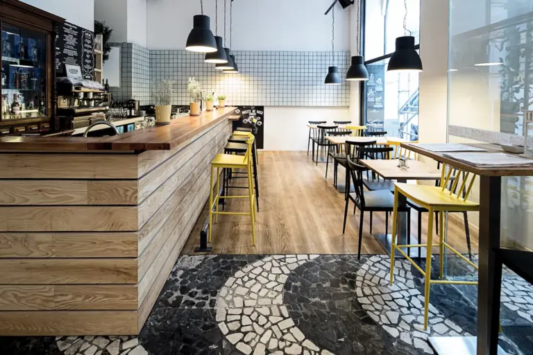 Modernes Café-Interieur mit Holztresen, gelben Hockern, schwarzen Pendelleuchten, hellem Holzfußboden, gefliester Wand und Mosaikfliesenboden in der Nähe des Eingangs. Tische und Stühle sind ordentlich angeordnet, mit großen Fenstern im Hintergrund.