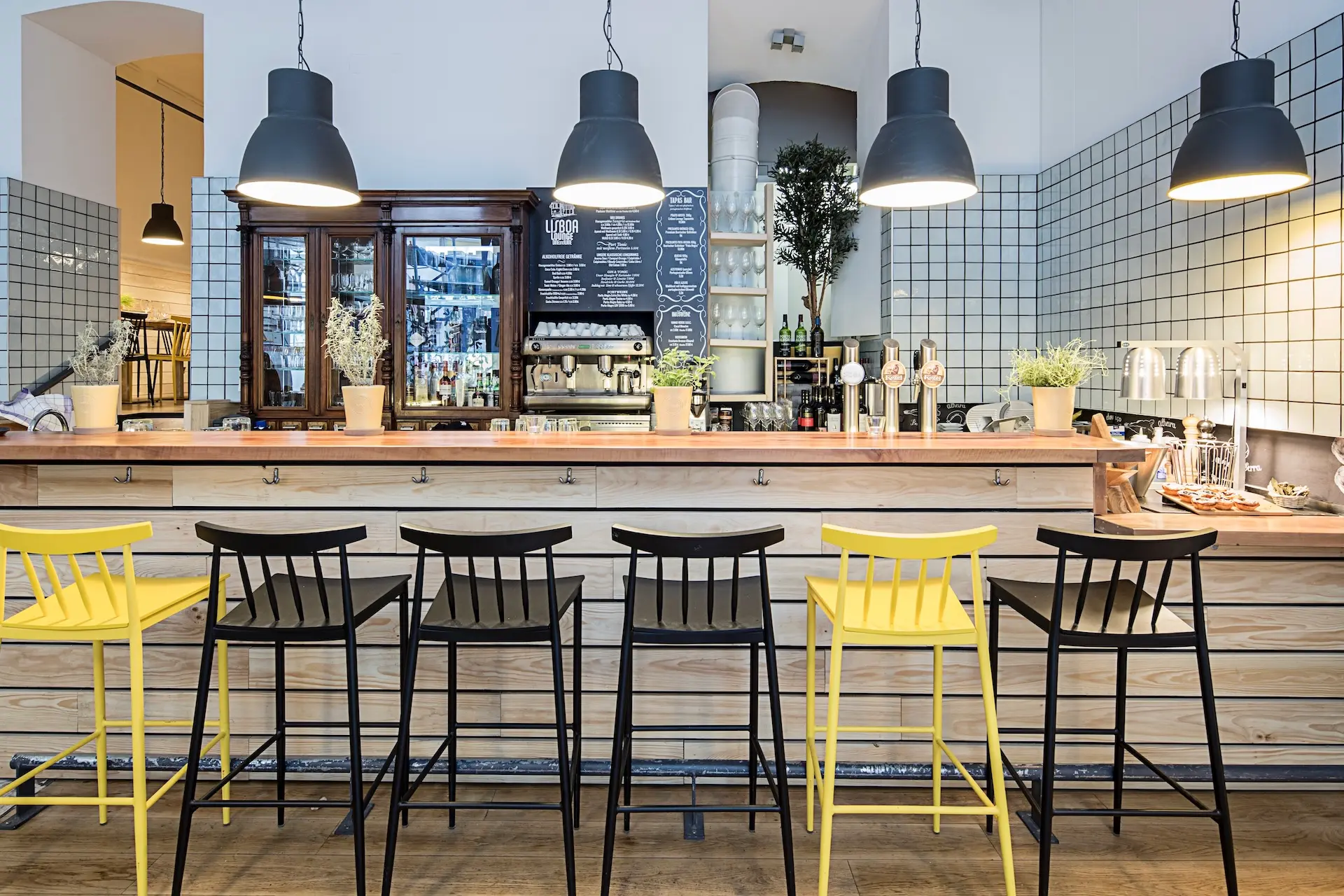Modernes Café-Interieur mit einem Holztresen, fünf Barhockern (drei schwarze, zwei gelbe), Topfpflanzen, Hängeleuchten und einer gefliesten Wand mit Regalen und einer Speisekarte dahinter.