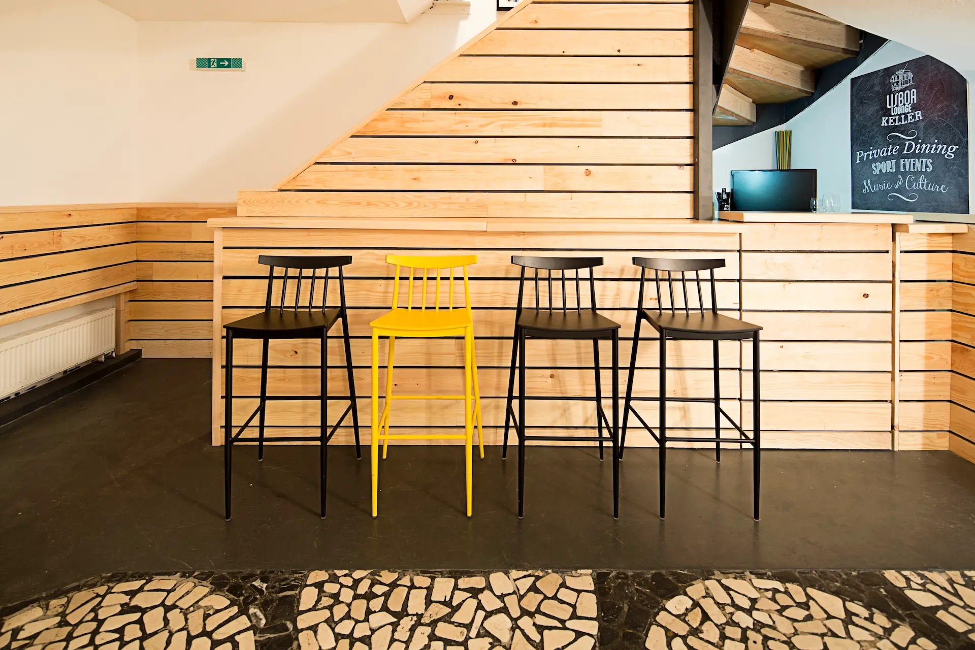 Vier hohe Stühle stehen vor einer modernen Holztheke, drei sind schwarz und einer in der Mitte ist gelb. Der Hintergrund besteht aus einer hellen Holzvertäfelung und einem geometrisch gefliesten Boden.
