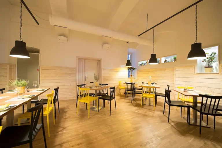 Ein modernes, minimalistisches Restaurant mit Holzböden, hellen Paneelwänden, gelben und schwarzen Stühlen, Holztischen zum Essen, Topfpflanzen und schwarzen Pendelleuchten an der Decke.